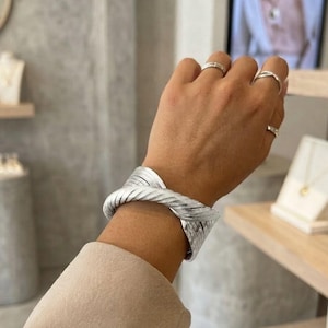 Peut inclure: Un large bracelet manchette argenté présentant un design torsadé à plusieurs brins avec une finition métallique. Le bracelet est porté au poignet, accompagné de deux bagues argentées simples sur l'index et le majeur de la même main.