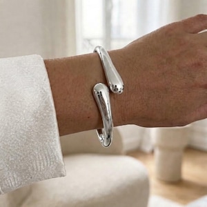 Peut inclure: Un bracelet manchette en métal argenté poli présentant un design ouvert avec deux extrémités lisses, arrondies et en forme de goutte. Le bracelet possède une finition métallique réfléchissante et une structure solide et incurvée.