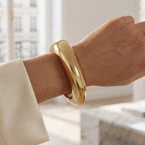 Peut inclure: Un bracelet manchette épais en métal doré poli avec une surface lisse, arrondie et réfléchissante. Ce jonc présente un design incurvé et substantiel qui entoure le poignet, mettant en valeur une finition métallique brillante et une structure solide.