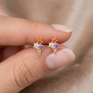 Peut inclure: Une paire de boucles d'oreilles à clous en or, chacune composée d'un groupe de trois pierres précieuses rondes orange, blanches et violettes, disposées en triangle se terminant en pointe, montées sur une structure en métal doré poli.
