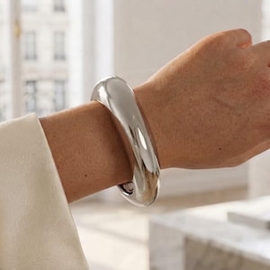 Op de afbeelding: Een dikke, gepolijste zilverkleurige armband met een glad, afgerond en doorlopend oppervlak. De armband heeft een hoogglanzende metalen afwerking die licht reflecteert, wat zorgt voor een gedurfde, solide en gebogen vorm rond de pols.