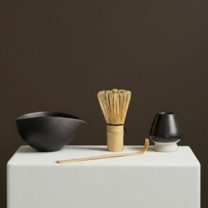 Könnte beinhalten: Eine schwarze Matcha-Schale aus Keramik, ein Schneebesen, ein Löffel und ein Chawan-Ständer sind auf einer weißen Oberfläche angeordnet. Der Schneebesen ist aus Bambus und der Chawan-Ständer ist schwarz mit weißem Sockel. Der Hintergrund ist ein dunkles Braun.