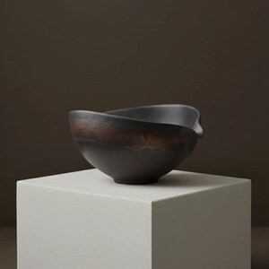 May include: A dark brown ceramic bowl with a unique, asymmetrical rim. The bowl has a textured, earthy appearance with a band of lighter brown near the top. It sits on a white, geometric pedestal, creating a minimalist aesthetic.