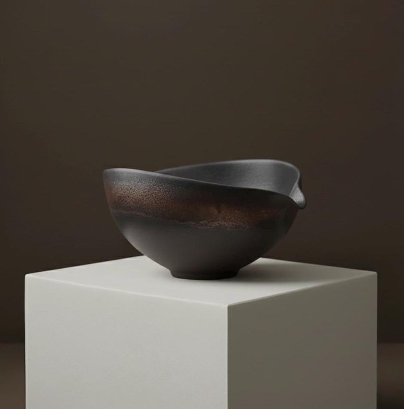 May include: A dark brown ceramic bowl with a unique, asymmetrical rim. The bowl has a textured, earthy appearance with a band of lighter brown near the top. It sits on a white, geometric pedestal, creating a minimalist aesthetic.