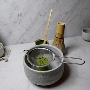 May include: A matcha tea preparation setup featuring a gray ceramic bowl with a stainless steel strainer holding green matcha powder. A bamboo whisk, a bamboo scoop, and a matcha tin are also present, all on a white marble surface.