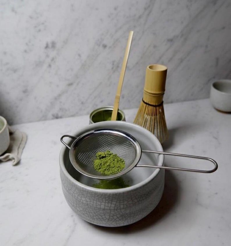 May include: A matcha tea preparation setup featuring a gray ceramic bowl with a stainless steel strainer holding green matcha powder. A bamboo whisk, a bamboo scoop, and a matcha tin are also present, all on a white marble surface.