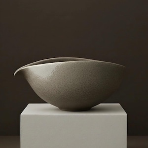 May include: A speckled, gray ceramic bowl with a curved spout. The bowl has a smooth, glossy finish and sits on a white, square pedestal against a dark brown background. The bowl's design is minimalist and modern.
