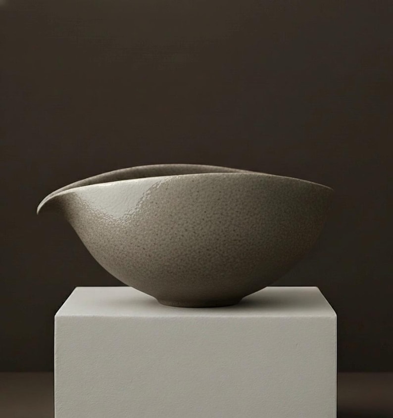 May include: A speckled, gray ceramic bowl with a curved spout. The bowl has a smooth, glossy finish and sits on a white, square pedestal against a dark brown background. The bowl's design is minimalist and modern.