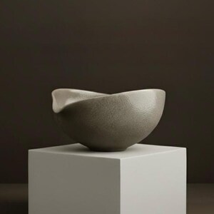 May include: A gray ceramic bowl with a unique, asymmetrical design. The bowl has a textured surface and sits on a white square pedestal against a dark brown background. The bowl's rim curves in an elegant, organic shape.