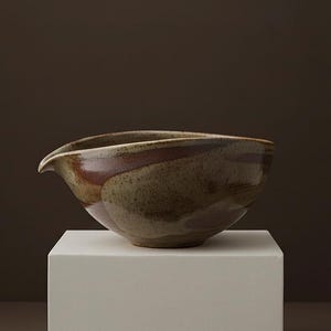 May include: A handcrafted ceramic bowl with a spout, featuring earthy tones of brown, tan, and green. The bowl is placed on a white pedestal, showcasing its unique shape and design. This piece is ideal for serving or as a decorative accent.