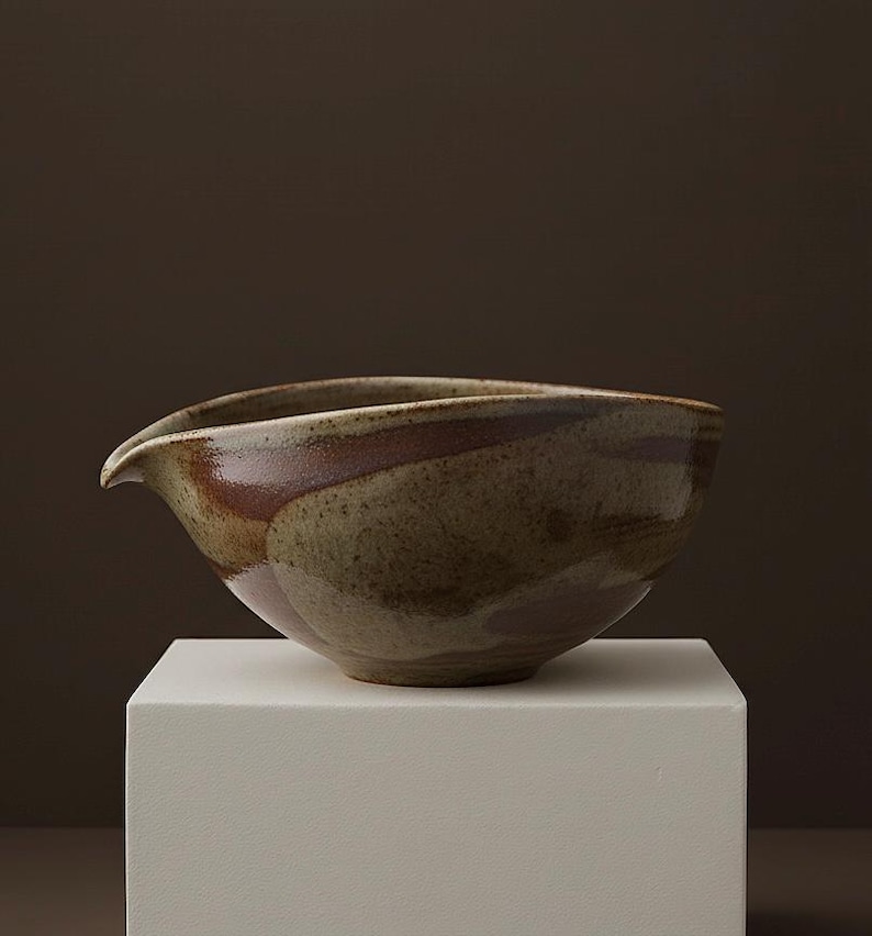 May include: A handcrafted ceramic bowl with a spout, featuring earthy tones of brown, tan, and green. The bowl is placed on a white pedestal, showcasing its unique shape and design. This piece is ideal for serving or as a decorative accent.