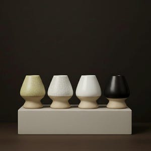 Peut inclure: Quatre fouets à matcha en céramique de différentes couleurs : vert pâle, blanc cassé avec un fini craquelé, blanc uni et noir. Chaque fouet a une base beige clair et repose sur un socle rectangulaire gris clair. Le fond est marron foncé.