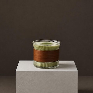 May include: A clear, vertically ribbed glass cup filled with a light green matcha latte, featuring a smooth wooden sleeve wrapped around the center. The glass has a wide, open top and a cylindrical shape, showcasing the textured surface of the glass.