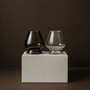 Könnte beinhalten: Zwei Trinkgläser mit rundem Boden und breiter Öffnung stehen auf einem weißen Block. Ein Glas ist klar, das andere dunkelgrau. Der Hintergrund ist dunkelbraun.