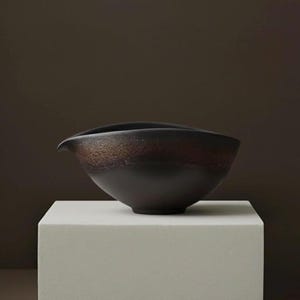 May include: A dark brown ceramic bowl with a unique spout design. The bowl has a textured, earthy appearance with a gradient of colors from dark brown to a lighter, reddish-brown. It sits on a white, square pedestal against a dark brown backdrop.