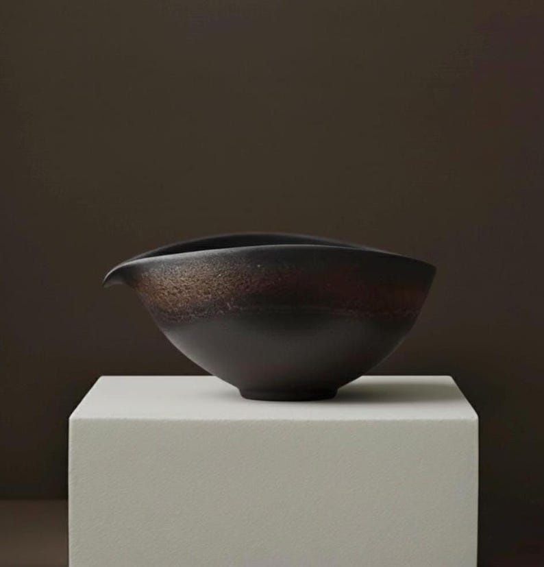 May include: A dark brown ceramic bowl with a unique spout design. The bowl has a textured, earthy appearance with a gradient of colors from dark brown to a lighter, reddish-brown. It sits on a white, square pedestal against a dark brown backdrop.