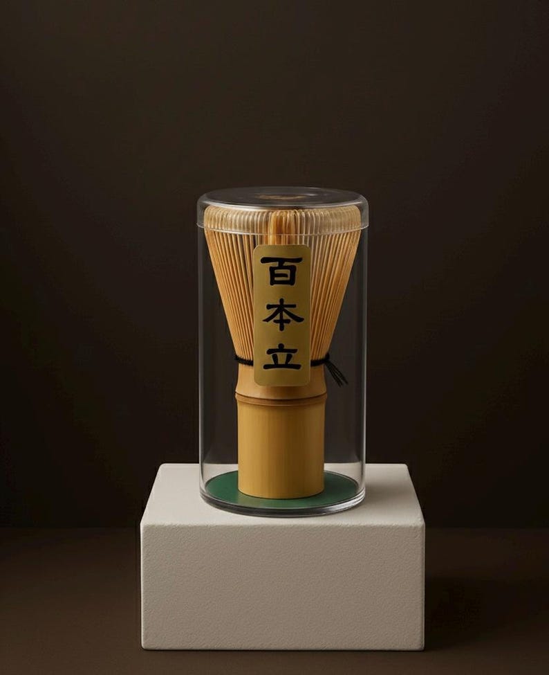 May include: A bamboo matcha whisk, or chasen, inside a clear cylindrical container. The whisk is light brown with a bamboo handle and a tag with Japanese text. The container sits on a white block against a dark background.