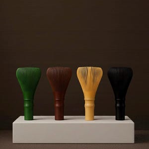 May include: Four matcha whisks in a row, each in a different colour. The whisks are green, brown, yellow, and black. They are displayed on a white rectangular platform against a dark brown background. The whisks have a bamboo-like handle.