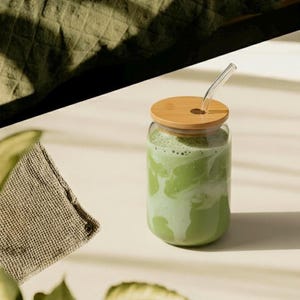 Peut inclure: Un verre transparent rempli d'une boisson verte, surmont&eacute; d'un couvercle en bois et d'une paille en verre transparent. Un petit tissu beige et des feuilles vertes sont au premier plan. La boisson semble &ecirc;tre un matcha latte.