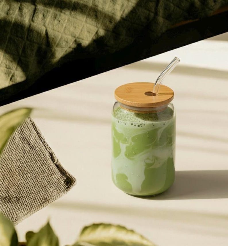Peut inclure: Un verre transparent rempli d'une boisson verte, surmont&eacute; d'un couvercle en bois et d'une paille en verre transparent. Un petit tissu beige et des feuilles vertes sont au premier plan. La boisson semble &ecirc;tre un matcha latte.