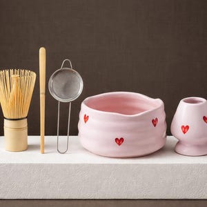 Peut inclure: Un service à thé matcha sur une surface blanche. L'ensemble comprend un bol en céramique rose et un petit vase, chacun décoré de cœurs rouges. Un fouet en bambou, une cuillère en bambou et un tamis en métal sont également inclus.