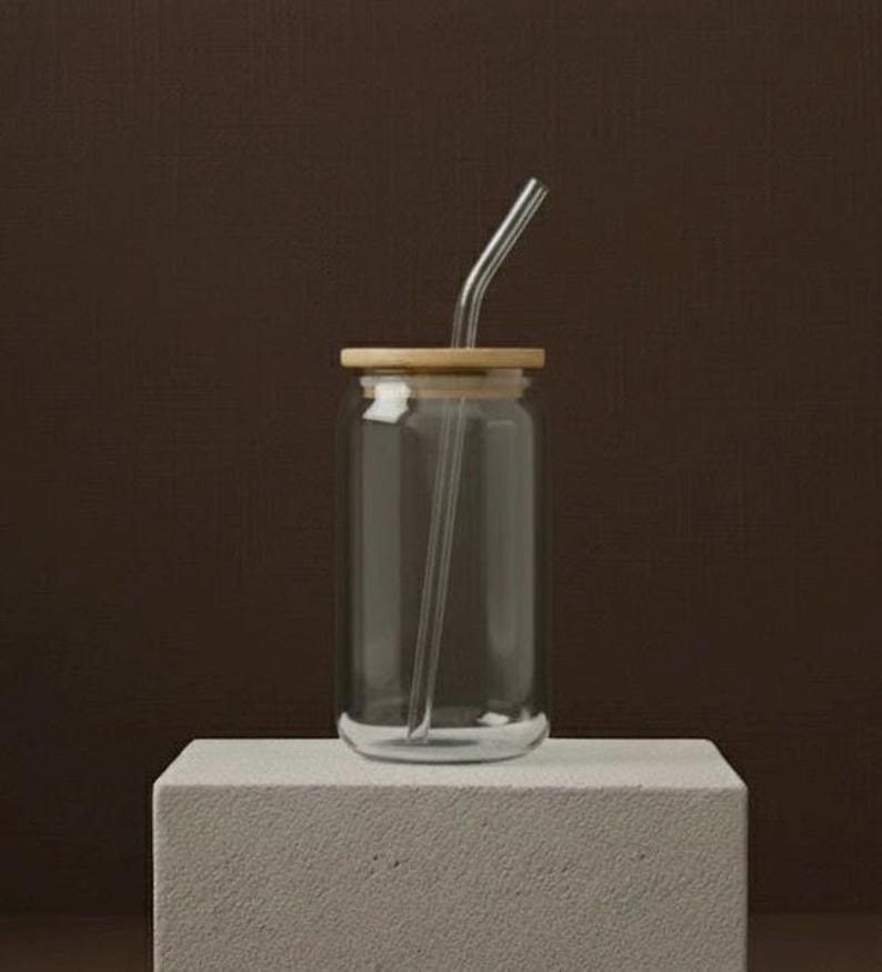 Peut inclure: Un verre &agrave; boire transparent en forme de canette avec un couvercle en bambou et une paille en verre transparent. Le verre repose sur un bloc rectangulaire blanc sur fond marron. La paille est courb&eacute;e en haut.