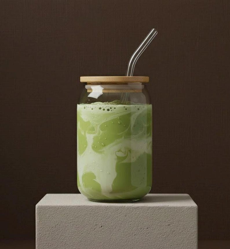 Peut inclure: Un verre transparent rempli d'une boisson verte et blanche, surmont&eacute; d'un couvercle en bambou et d'une paille en verre transparent. Le verre est pos&eacute; sur un bloc rectangulaire blanc sur fond marron.