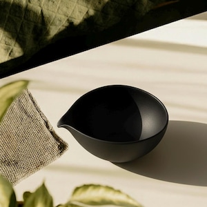 May include: A matte black ceramic bowl with a spout, ideal for serving sauces or dips. The bowl has a smooth, rounded shape and sits on a light-colored surface. A small, textured, tan cloth is next to the bowl. The image is lit by natural light.