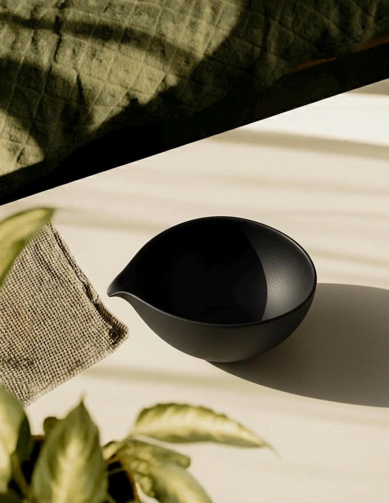 May include: A matte black ceramic bowl with a spout, ideal for serving sauces or dips. The bowl has a smooth, rounded shape and sits on a light-colored surface. A small, textured, tan cloth is next to the bowl. The image is lit by natural light.