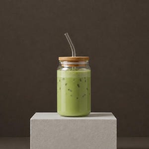 Op de afbeelding: Een doorzichtig glas gevuld met een lichtgroene matcha latte en ijsblokjes. Het glas is voorzien van een rond bamboe deksel met een gat in het midden en een gebogen, doorzichtig glazen rietje dat boven uit de drankhouder steekt.