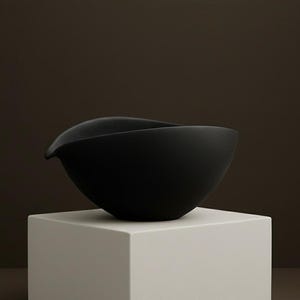 May include: A matte black ceramic bowl with a unique, asymmetrical design. The bowl has a smooth, curved shape with a small spout on one side. It is displayed on a white, square pedestal against a dark brown background.