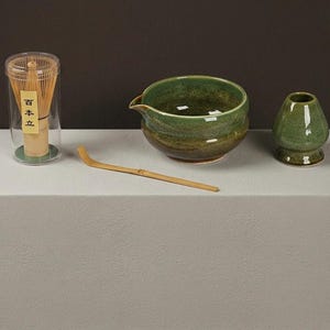 Peut inclure: Un ensemble de thé matcha comprenant un fouet en bambou dans un étui transparent, un bol en céramique vert avec un bec verseur, un petit bol à thé en céramique vert et une cuillère en bambou. Les articles sont disposés sur une surface blanche.