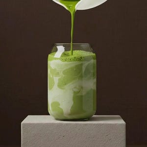 Peut inclure: Un verre transparent rempli d'une boisson verte et de tourbillons blancs, avec plus de liquide vert vers&eacute; d'un pichet blanc. La boisson est sur un bloc blanc sur fond marron.