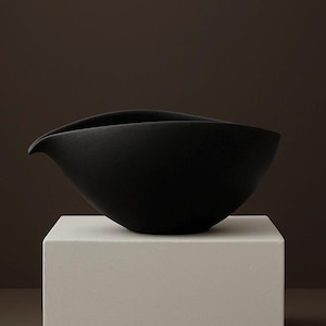 Könnte beinhalten: Eine matte, schwarze Keramikschale mit einem einzigartigen, asymmetrischen Design. Die Schale hat einen geschwungenen Rand und eine auslaufartige Verlängerung an einer Seite. Sie steht auf einem weißen, quadratischen Sockel vor einem dunkelbraunen Hintergrund.