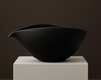 May include: A matte black ceramic bowl with a unique, asymmetrical design. The bowl has a curved rim and a spout-like extension on one side. It sits on a white, square pedestal against a dark brown background.