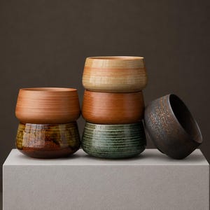 Op de afbeelding: Een verzameling van vijf keramische kommen gerangschikt in een stapel en een enkele schuine kom. De kommen hebben gevarieerde afwerkingen, waaronder glad lichtbruin, mat terracotta, glanzend donkergroen met horizontale ribbels en een donker gespikkeld antraciet.