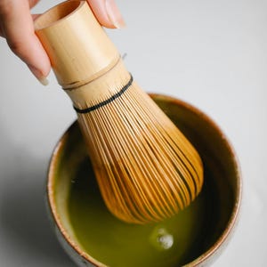 May include: A bamboo matcha whisk, or chasen, is dipped into a ceramic bowl of vibrant green matcha tea. The bowl has a speckled, earthy design. The whisk is made of thin bamboo tines, and is used to mix matcha powder with hot water.