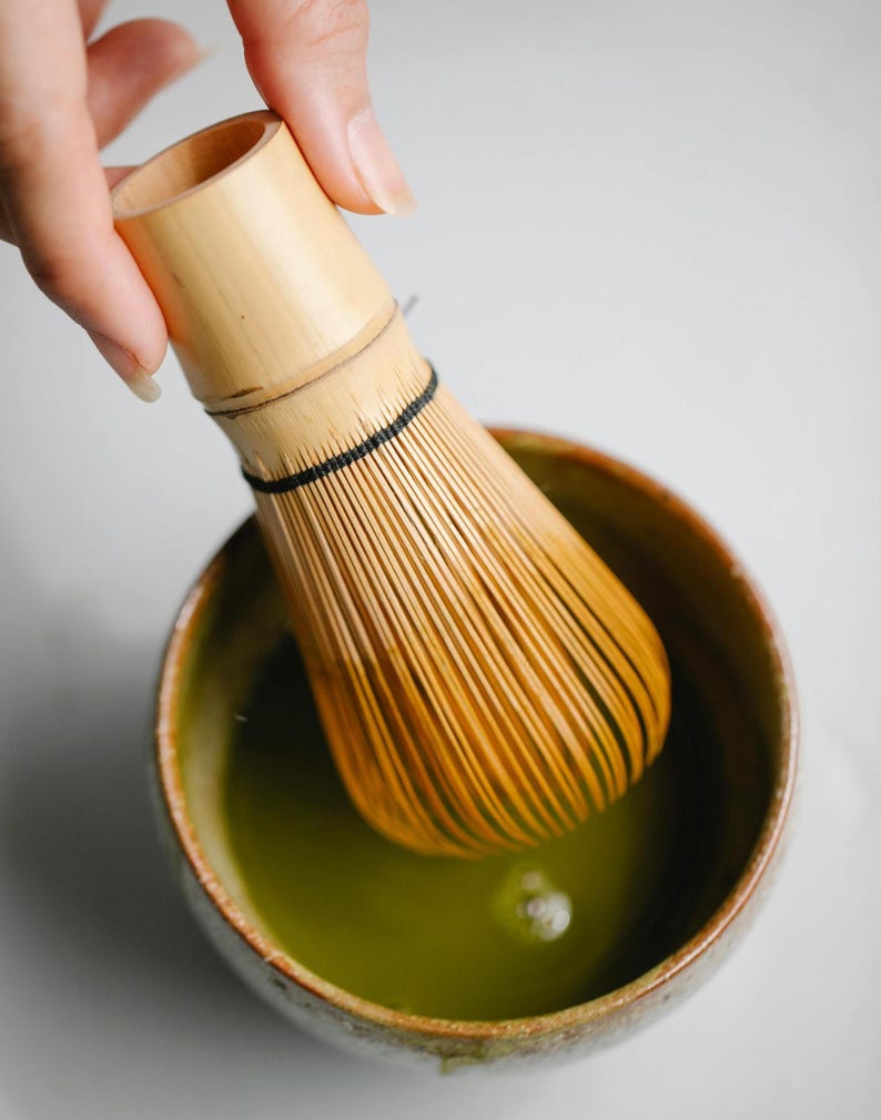 May include: A bamboo matcha whisk, or chasen, is dipped into a ceramic bowl of vibrant green matcha tea. The bowl has a speckled, earthy design. The whisk is made of thin bamboo tines, and is used to mix matcha powder with hot water.