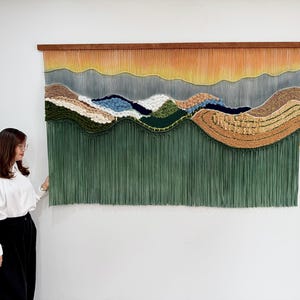 以下が含まれることがあります： 風景デザインの大きなテクスチャードウォールハンギング。このアートワークは、オレンジ、イエロー、ブルー、グリーン、ブラウンなどの色のグラデーションが特徴です。木製のトップと、下部に長いグリーンのフリンジが付いています。