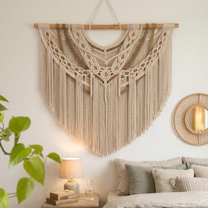 May include: A large, beige macrame wall hanging with intricate woven patterns and fringe. The textile is suspended from a wooden dowel, creating a bohemian aesthetic. The wall decor is a focal point in a bedroom setting.