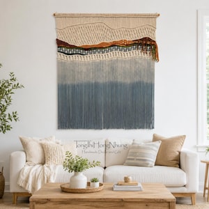 May include: A large macrame wall hanging featuring a landscape design with cream, brown, green, and blue fiber layers. The top section has intricate knotting, while the bottom consists of long, straight, blue-toned fringe hanging from a wooden rod.