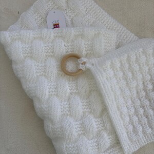 Könnte beinhalten: Weiße gestrickte Babydecke mit einem daran befestigten Holzring. Die Decke hat ein strukturiertes Muster und ist in der Hälfte gefaltet. Ein kleines Etikett mit dem Text "Cofi's" ist an der Decke befestigt.