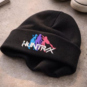 May include: Black knit beanie with the word "HUNTRAX" embroidered in white, with a colorful graphic of three figures. The figures are in blue, purple, and pink. The beanie is folded over at the brim.
