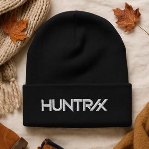 May include: A black beanie with the white embroidered text "HUNTR/X" on the front. The beanie is displayed on a neutral-toned surface with a scarf, boots, and a fall leaf.