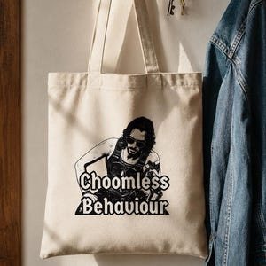 Puede incluir: Un bolso tote beige con un gráfico negro de una persona y el texto "Choomless Behaviour". El bolso cuelga de un gancho de pared junto a una chaqueta vaquera. El bolso tiene dos asas.