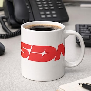 Puede incluir: Una taza de café de cerámica blanca con las letras rojas "SDN" impresas en el lateral. La taza está llena de café oscuro y está sobre un escritorio, junto a un bloc de notas, un bolígrafo y un ratón de ordenador.