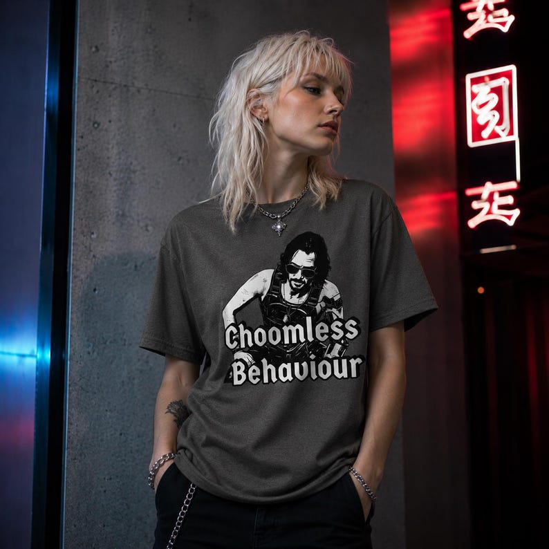 May include: Dark grey t-shirt with a graphic of a man wearing sunglasses and the words "Choomless Behaviour" in white. The shirt is worn by a person with blonde hair, a silver necklace, and a silver bracelet. The background includes a dark grey wall and a red neon sign.