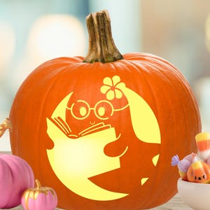 May include: A large orange pumpkin with a carved design of a crescent moon character wearing round glasses and a flower, holding an open book. The carving is illuminated from within, emitting a bright yellow light against the pumpkin skin.
