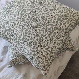 May include: Two square throw pillows featuring a dense, repeating botanical pattern of sage green leaves, stems, and small flowers printed on a white fabric background. The pillows are arranged overlapping each other, showcasing the intricate floral design.
