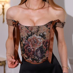 May include: A floral tapestry corset with brown ribbon ties and off-the-shoulder sleeves. The corset features a pattern of roses in shades of brown, red, and purple. The model is wearing a black skirt and a gold necklace.